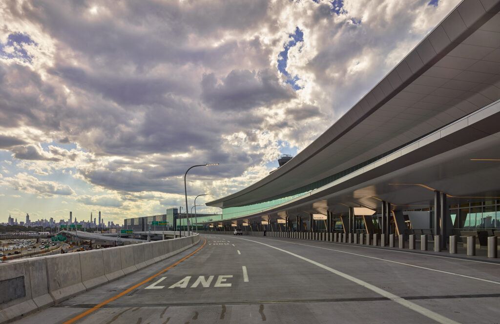 Airport dropoff/arrival lane in front of LaGuardia new terminal. No cars on the lane. City is visible in the background.