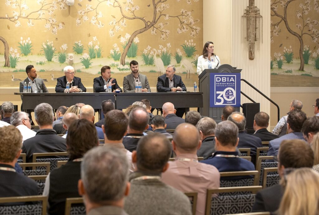 A crowd of people watching a woman at a lectern speaking as part of a panel presentation at a conference